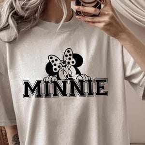 May include: A light beige t-shirt featuring a black and white graphic of Minnie Mouse peeking over the word "MINNIE" in bold, black letters. The design includes Minnie's iconic polka dot bow. The shirt has a crew neck and short sleeves.