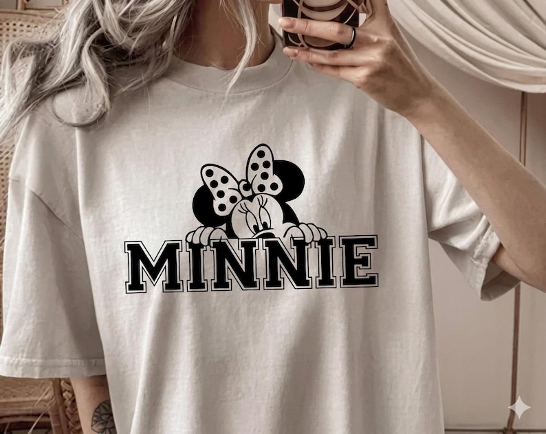 May include: A light beige t-shirt featuring a black and white graphic of Minnie Mouse peeking over the word "MINNIE" in bold, black letters. The design includes Minnie's iconic polka dot bow. The shirt has a crew neck and short sleeves.