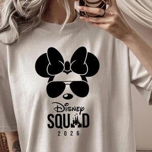May include: A light beige t-shirt featuring a black graphic of Minnie Mouse wearing sunglasses and a bow. Below the graphic, the text reads "Disney Squad 2026".