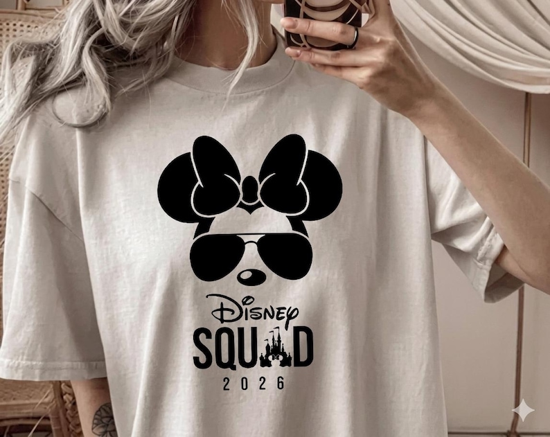 May include: A light beige t-shirt featuring a black graphic of Minnie Mouse wearing sunglasses and a bow. Below the graphic, the text reads "Disney Squad 2026".
