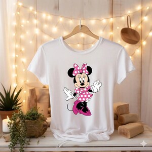 Puede incluir: Camiseta blanca con un dise&ntilde;o de Minnie Mouse. El personaje de dibujos animados lleva un vestido y un lazo de lunares rosas, con orejas y zapatos negros. El dise&ntilde;o est&aacute; centrado en la camiseta, que cuelga de una percha de madera.