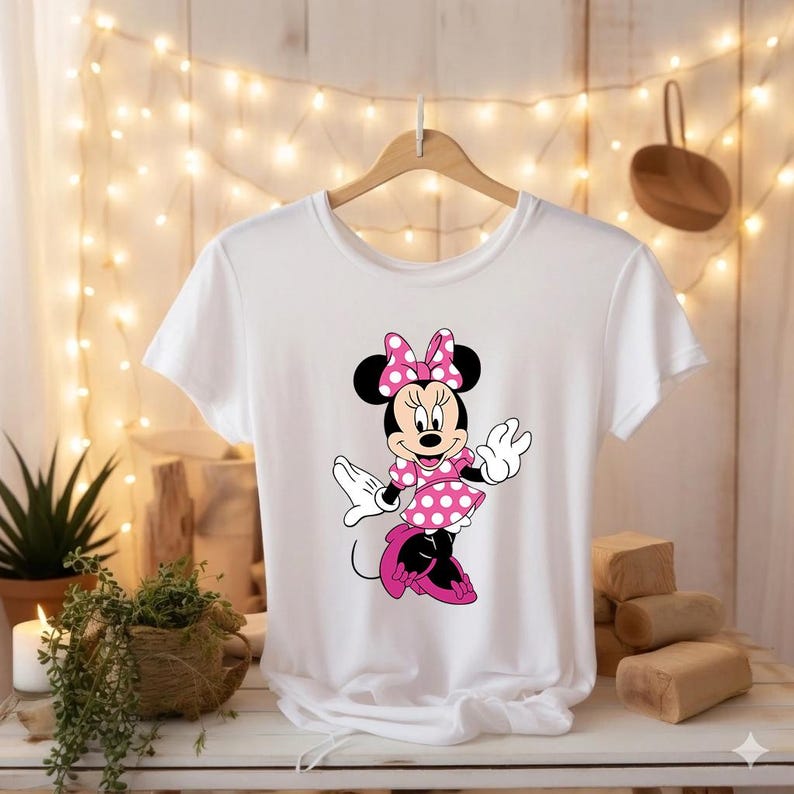 Puede incluir: Camiseta blanca con un dise&ntilde;o de Minnie Mouse. El personaje de dibujos animados lleva un vestido y un lazo de lunares rosas, con orejas y zapatos negros. El dise&ntilde;o est&aacute; centrado en la camiseta, que cuelga de una percha de madera.