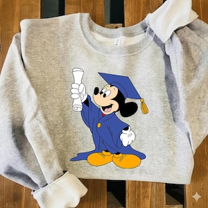 Puede incluir: Una sudadera gris con un dise&ntilde;o de Mickey Mouse con tem&aacute;tica de graduaci&oacute;n. Mickey est&aacute; representado con una toga y birrete azules, sosteniendo un diploma. La sudadera tiene pu&ntilde;os blancos y un cuello blanco.
