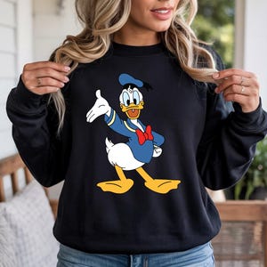 Puede incluir: Sudadera negra con un gr&aacute;fico colorido de Donald Duck con un atuendo de marinero. El personaje de dibujos animados lleva un traje de marinero azul, una pajarita roja y un gorro de marinero. La sudadera es de cuello redondo.