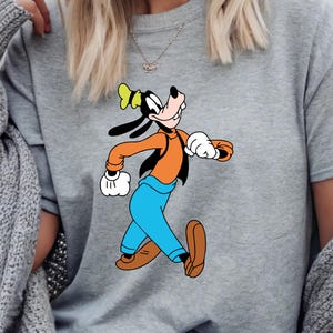 Puede incluir: Una sudadera gris con una imagen de dibujos animados de Goofy corriendo. Goofy lleva una camisa naranja, pantalones azules, zapatos marrones y un sombrero amarillo. La sudadera es de estilo casual.