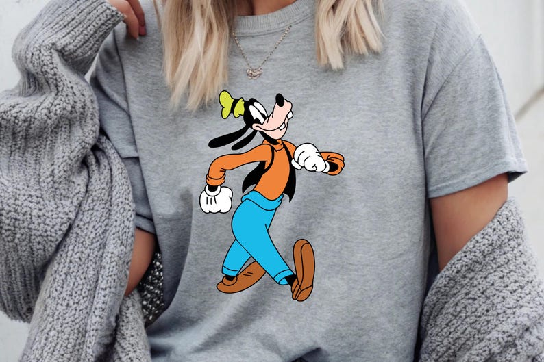 Puede incluir: Una sudadera gris con una imagen de dibujos animados de Goofy corriendo. Goofy lleva una camisa naranja, pantalones azules, zapatos marrones y un sombrero amarillo. La sudadera es de estilo casual.