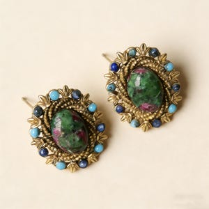 May include: A pair of vintage-style stud earrings. Each earring features an oval-shaped, green and red stone, surrounded by small blue and turquoise stones, set in a gold-toned metal frame.