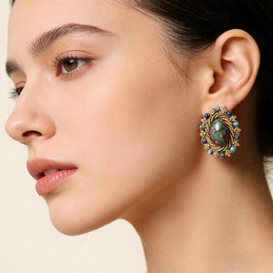 Peut inclure: Gros plan d'une boucle d'oreille dor&eacute;e avec une pierre centrale ovale aux teintes rouges et vertes. La boucle d'oreille est entour&eacute;e de petites pierres bleues et turquoise, et d'un motif filigran&eacute; dor&eacute;.