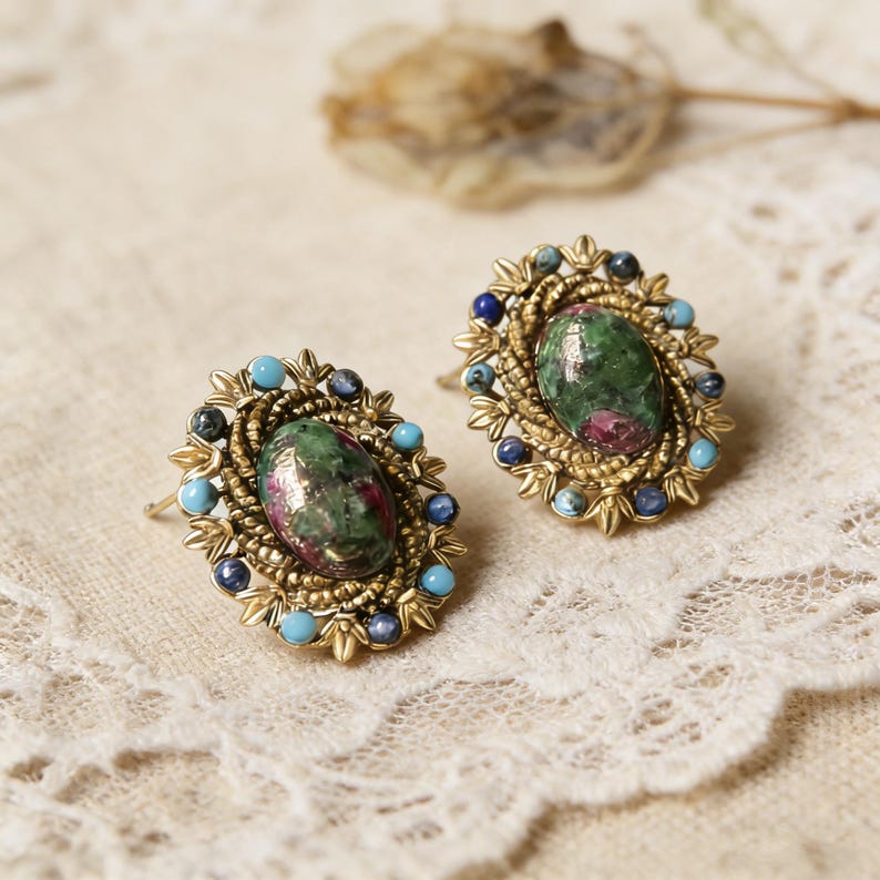 Peut inclure: Une paire de boucles d'oreilles de style vintage. Chaque boucle d'oreille pr&eacute;sente une pierre pr&eacute;cieuse ovale verte et rouge au centre, entour&eacute;e d'un cadre en filigrane dor&eacute; et orn&eacute;e de petites perles bleues et turquoise. Les boucles d'oreilles sont pos&eacute;es sur un fond en dentelle blanc cass&eacute;.