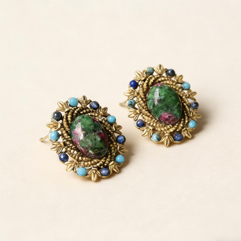 Peut inclure: Paire de boucles d'oreilles ovales. Chaque boucle pr&eacute;sente une pierre verte centrale avec des inclusions rouges, entour&eacute;e d'un cadre dor&eacute; et orn&eacute;e de perles bleues et turquoise. Les boucles d'oreilles ont un style vintage.