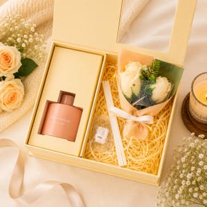 May include: A gift box containing a peach-colored bottle labeled HOME FRAGRANCE, a small bouquet of two cream-colored roses wrapped in peach paper with a white ribbon, a white reed diffuser stick, and a small white plastic component nestled in straw.
