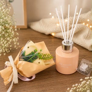 May include: A peach-colored cylindrical glass home fragrance diffuser with five white reed sticks and a silver collar, accompanied by a small bouquet of artificial flowers wrapped in peach paper with a white ribbon and a small clear bag of fairy lights.