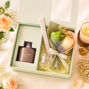 May include: A light green gift box containing a brown bottle labelled HOME FRAGRANCE RELAX AT HOME, a small bouquet of green and cream roses wrapped in netting with a ribbon, and a set of light-coloured reed diffuser sticks nestled in straw.