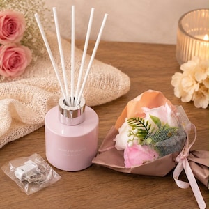 May include: A light pink cylindrical home fragrance diffuser with a silver collar and five white reed sticks. The bottle features the text HOME FRAGRANCE printed on the front. A small plastic bag with a white clip and a small bouquet of pink flowers.