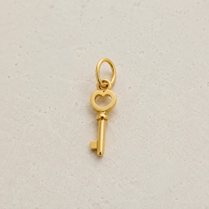 May include: A gold-toned metal charm in the shape of a key with a heart-shaped bow at the top. The charm features a circular jump ring attached to the top of the heart, a slender cylindrical shaft, and a rectangular bit at the bottom end.