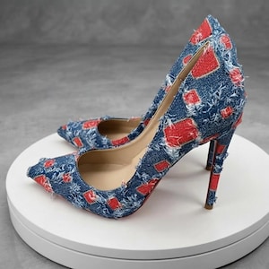 May include: A pair of pointed-toe high heel pumps crafted from blue denim fabric with a frayed, distressed texture. The shoes feature a repeating pattern of small, rectangular red patches throughout the exterior and a slim, high stiletto heel.