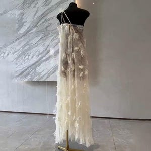 Puede incluir: Un vestido transparente de color blanco roto con delicados apliques florales. El vestido presenta una sola correa para el hombro y un dise&ntilde;o largo y fluido. Se exhibe en un maniqu&iacute; negro sobre un fondo neutro, mostrando su cualidad et&eacute;rea.