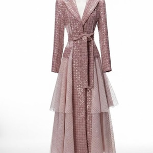May include: A long, pink coat with a matching belt and a tiered skirt. The coat features a notched collar and long sleeves. The skirt has a dotted overlay. The coat is displayed on a mannequin.