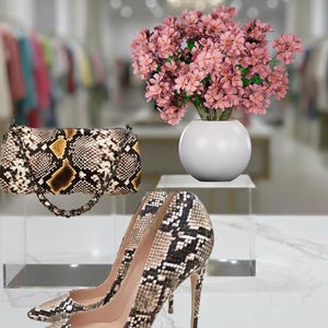 May include: A fashion-forward display featuring a snakeskin-patterned handbag and high heels. A white vase holds pink flowers, adding a touch of elegance. The shoes have pointed toes and stiletto heels. The bag has a chain strap.