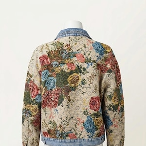 May include: A denim jacket with a floral pattern in shades of red, blue, yellow, and green. The jacket has a light-wash denim collar and cuffs. The floral design covers the entire back of the jacket.