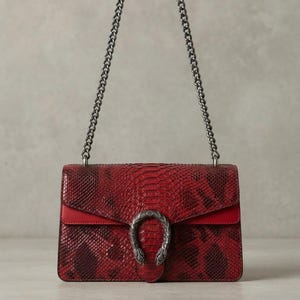 May include: A rectangular shoulder bag featuring a red and black snakeskin-textured pattern. The bag includes a silver-toned metal chain strap and a prominent silver-toned horseshoe-shaped clasp with two animal head details at the center front.