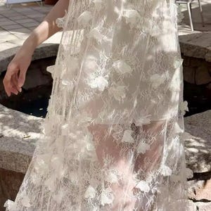 Puede incluir: Un vestido blanco largo con una delicada capa de encaje. El vestido presenta aplicaciones florales tridimensionales dispersas sobre la tela. El vestido parece estar hecho de un material ligero, adecuado para climas c&aacute;lidos u ocasiones especiales.