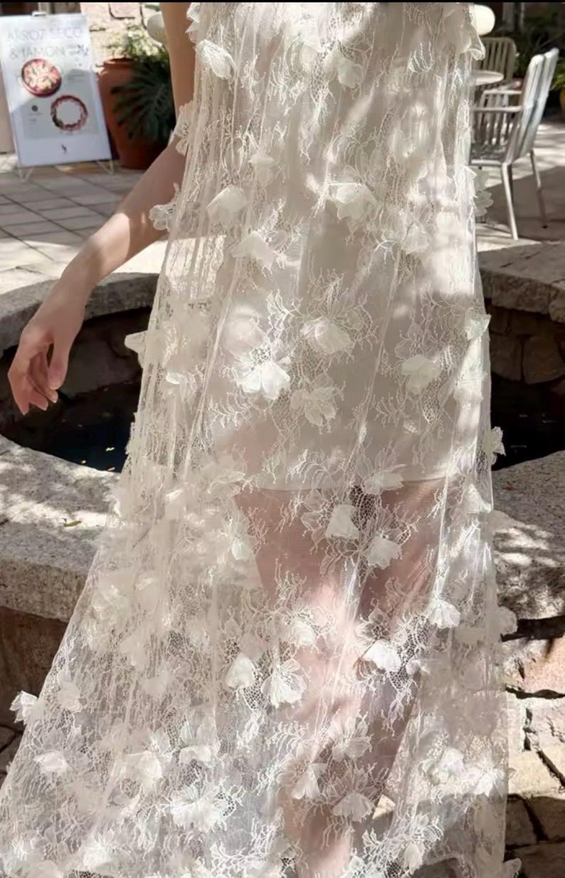 Puede incluir: Un vestido blanco largo con una delicada capa de encaje. El vestido presenta aplicaciones florales tridimensionales dispersas sobre la tela. El vestido parece estar hecho de un material ligero, adecuado para climas c&aacute;lidos u ocasiones especiales.