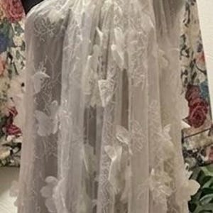 Puede incluir: Un vestido blanco de un solo hombro, hecho de tela transparente, adornado con aplicaciones florales tridimensionales. El vestido se exhibe en un maniqu&iacute; negro, con una chaqueta con estampado floral visible en el fondo. El vestido parece ser largo.