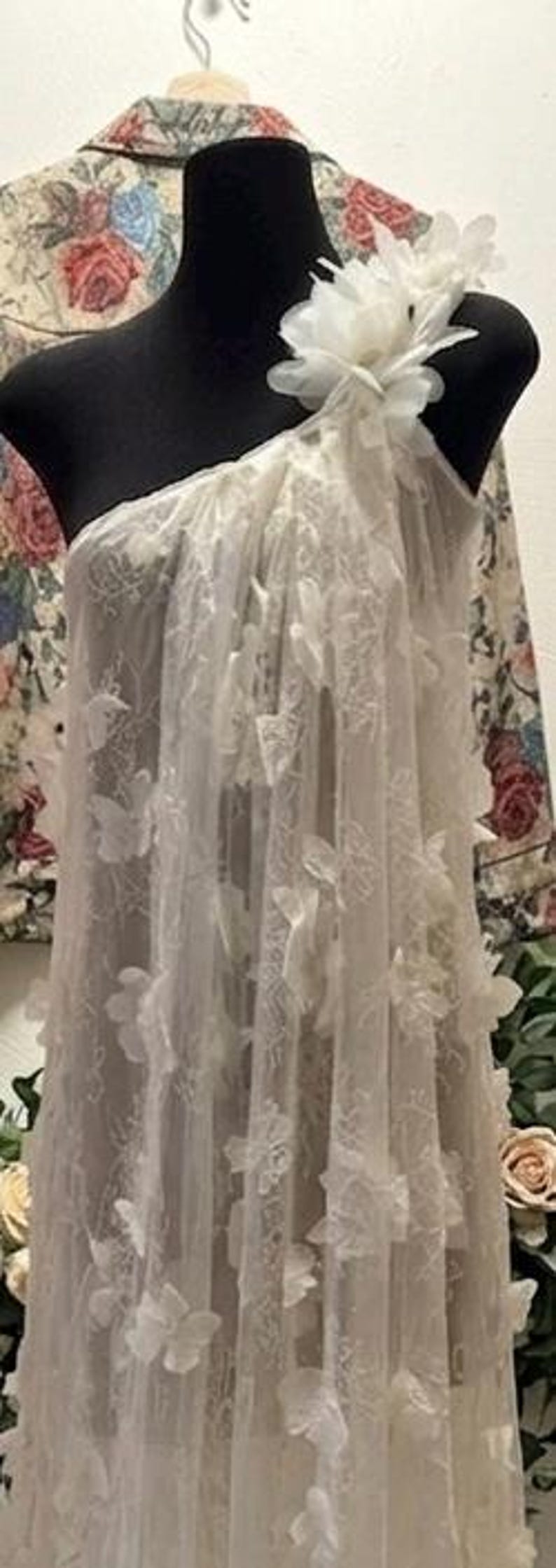 Puede incluir: Un vestido blanco de un solo hombro, hecho de tela transparente, adornado con aplicaciones florales tridimensionales. El vestido se exhibe en un maniqu&iacute; negro, con una chaqueta con estampado floral visible en el fondo. El vestido parece ser largo.