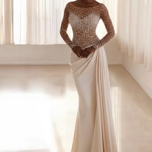 Puede incluir: Vestido largo color marfil con corpiño transparente con cuentas y mangas largas. La falda cae elegantemente hasta el suelo. Adecuado para eventos formales, como bodas o galas.