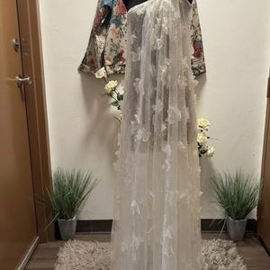 Puede incluir: Un vestido blanco de un solo hombro sobre un maniqu&iacute; negro. El vestido est&aacute; hecho de tela transparente y adornado con aplicaciones florales en 3D. Una chaqueta floral cuelga en el fondo. El vestido se exhibe sobre una alfombra de tonos neutros.