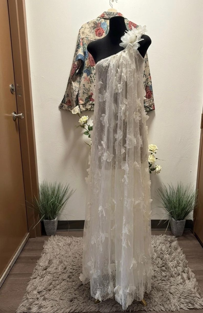 Puede incluir: Un vestido blanco de un solo hombro sobre un maniqu&iacute; negro. El vestido est&aacute; hecho de tela transparente y adornado con aplicaciones florales en 3D. Una chaqueta floral cuelga en el fondo. El vestido se exhibe sobre una alfombra de tonos neutros.