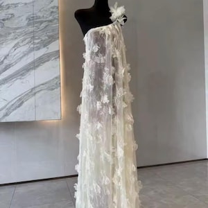 Puede incluir: Vestido blanco de un solo hombro sobre un maniqu&iacute; negro. El vestido est&aacute; hecho de tela transparente con aplicaciones florales. Una gran flor adorna el hombro. El vestido largo tiene una silueta fluida, ideal para eventos formales.