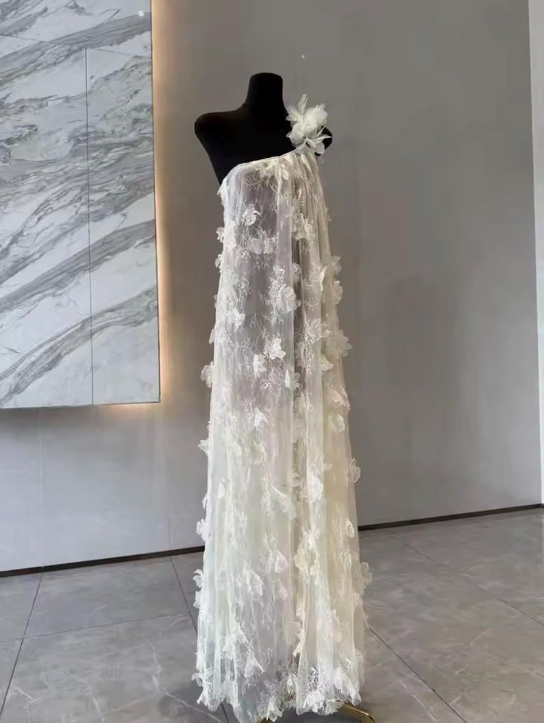 Puede incluir: Vestido blanco de un solo hombro sobre un maniqu&iacute; negro. El vestido est&aacute; hecho de tela transparente con aplicaciones florales. Una gran flor adorna el hombro. El vestido largo tiene una silueta fluida, ideal para eventos formales.