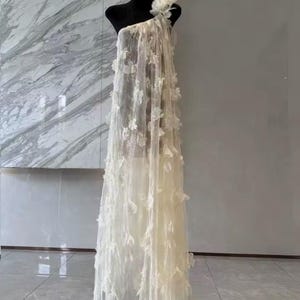 Puede incluir: Un vestido crema de un solo hombro sobre un maniqu&iacute; negro. El vestido est&aacute; hecho de tela transparente y adornado con delicadas aplicaciones florales tridimensionales. El vestido cae hasta el suelo, mostrando una silueta fluida. Una gran flor adorna el hombro.