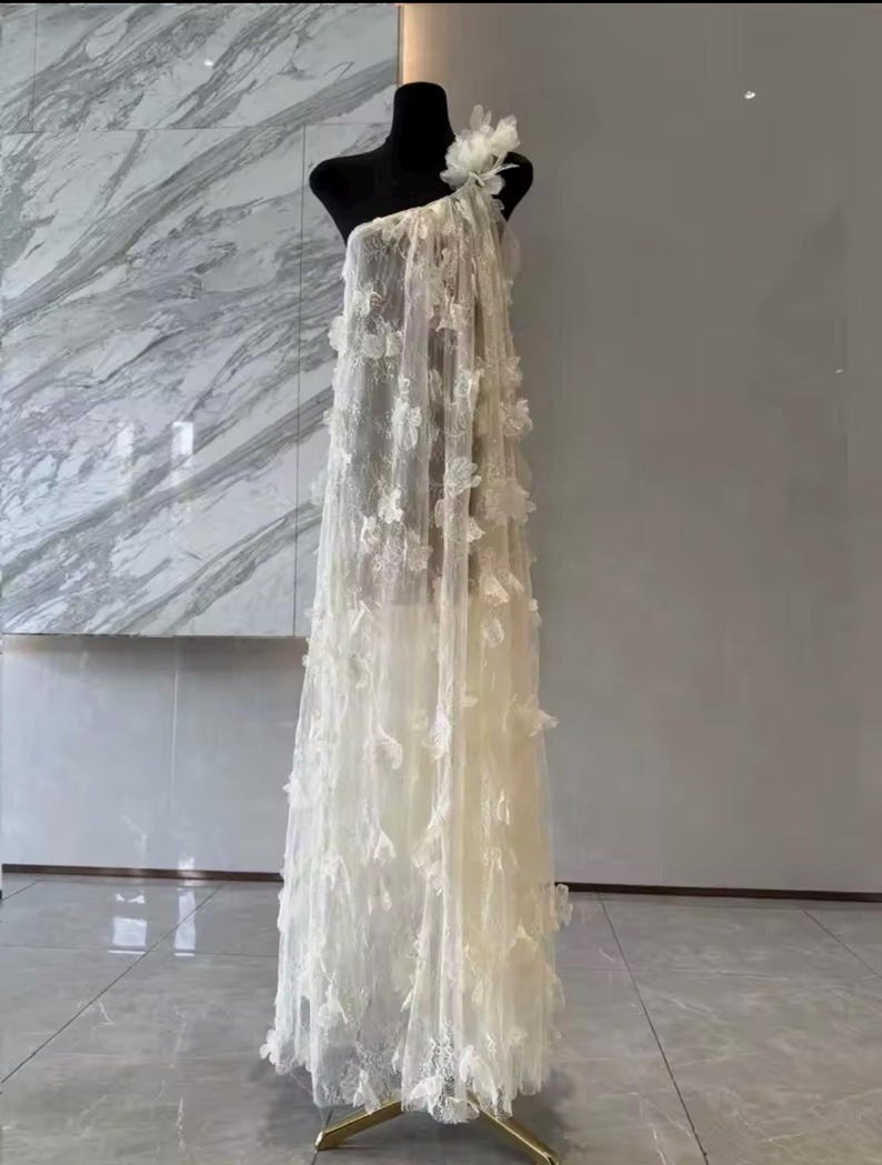 Puede incluir: Un vestido crema de un solo hombro sobre un maniqu&iacute; negro. El vestido est&aacute; hecho de tela transparente y adornado con delicadas aplicaciones florales tridimensionales. El vestido cae hasta el suelo, mostrando una silueta fluida. Una gran flor adorna el hombro.