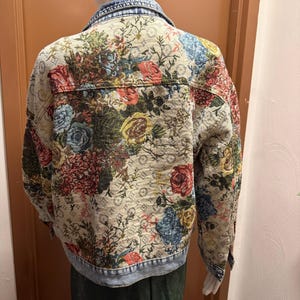 May include: A denim jacket with a floral tapestry design. The jacket features a light blue denim collar and cuffs, with a cream-colored body covered in a colorful floral pattern. The flowers include red, blue, and yellow blooms.