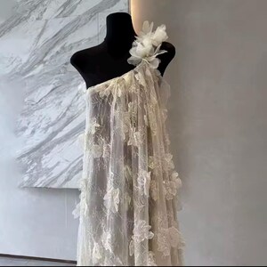 Puede incluir: Vestido marfil de un solo hombro sobre un maniqu&iacute; negro. El vestido est&aacute; hecho de tela transparente y adornado con aplicaciones florales tridimensionales. Un gran adorno floral acent&uacute;a el hombro. El vestido est&aacute; dise&ntilde;ado para ocasiones formales.