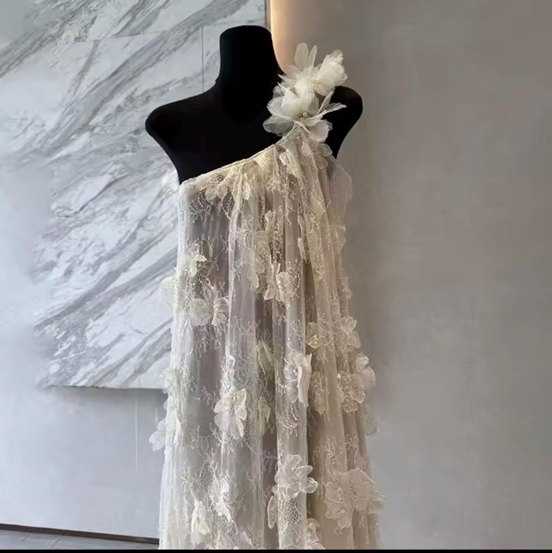 Puede incluir: Vestido marfil de un solo hombro sobre un maniqu&iacute; negro. El vestido est&aacute; hecho de tela transparente y adornado con aplicaciones florales tridimensionales. Un gran adorno floral acent&uacute;a el hombro. El vestido est&aacute; dise&ntilde;ado para ocasiones formales.