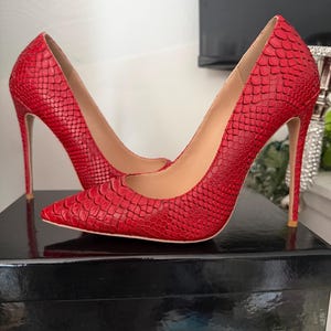 May include: A pair of red high-heeled shoes with a snakeskin pattern. The shoes have pointed toes and stiletto heels. The shoes are sitting on a black box.