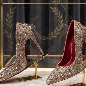 May include: A pair of gold stiletto pumps covered in shimmering crystals. The shoes feature a pointed toe, a high heel, and a deep red interior lining. Each shoe is displayed on a gold-toned metal stand, showcasing the exterior texture.