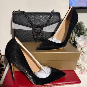 May include: A black leather handbag with a chain strap and a decorative clasp, paired with two black high-heeled pumps. The shoes have pointed toes and stiletto heels. The items are displayed on a shelf with a red book.