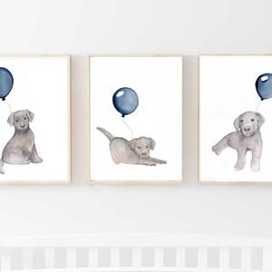 May include: Set of three framed watercolor prints featuring adorable gray puppies with blue balloons. Each print showcases a different puppy pose, all holding a balloon. The artwork is set against a white background, creating a charming nursery or children's room decor.