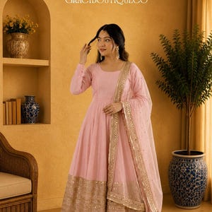 Op de afbeelding: Een lichtroze Anarkali-pak met gouden versieringen. De jurk met lange mouwen heeft een zwierige rok en een bijpassende dupatta met een gouden rand. De outfit wordt gedragen met bijpassende schoenen. De achtergrond omvat een geweven tapijt en decoratieve vazen.