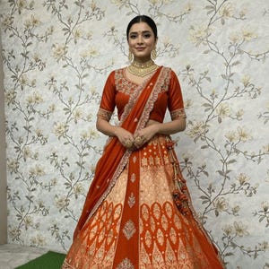 Könnte beinhalten: Eine Frau trägt einen traditionellen indischen Lehenga in Orange und Gold. Das Outfit umfasst eine kurzärmlige Bluse mit Stickerei, einen langen Rock mit aufwendigen Mustern und einen passenden Schal. Goldschmuck rundet den Look ab.
