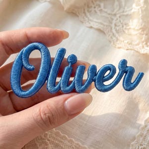 May include: A blue embroidered patch featuring the name Oliver in a cursive script font. The lettering is stitched with a dense, raised thread texture, creating a bold and defined appearance for the custom name design on a fabric base.