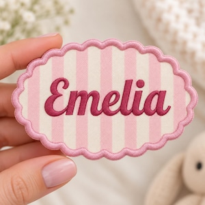 May include: An oval-shaped fabric patch with a scalloped edge, featuring a light pink and white vertical striped pattern. The name Emelia is embroidered in the center in a dark pink cursive script font with a thick border.