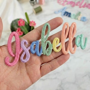 May include: A multi-coloured embroidered patch featuring the name Isabella in a cursive script font. Each letter is stitched in a different pastel thread colour, including pink, lavender, blue, light green, yellow, and peach, creating a vibrant effect.