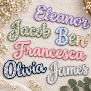May include: A collection of embroidered fabric name patches featuring the names Eleanor in purple floral, Jacob in green gingham, Ben in blue and tan gingham, Francesca in pink, Olivia in blue floral, and James in grey and white striped fabric.