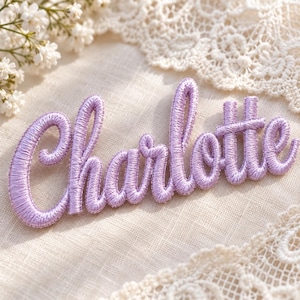 May include: A lavender-coloured embroidered patch featuring the name Charlotte in a cursive script font. The lettering is constructed with thick, raised satin stitching in a light purple thread, creating a textured and dimensional appearance.